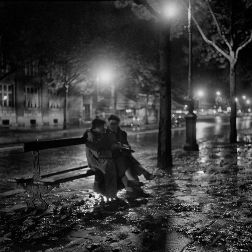 black and white photo of two people sitting together on a bench on a street late at night