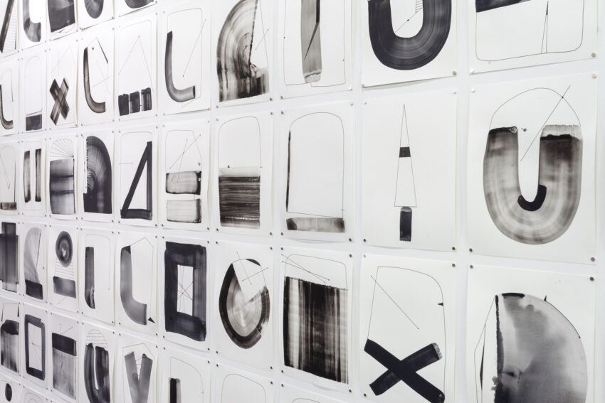 Installation of many peices of paper, each with black and gray shapes drawn on them.