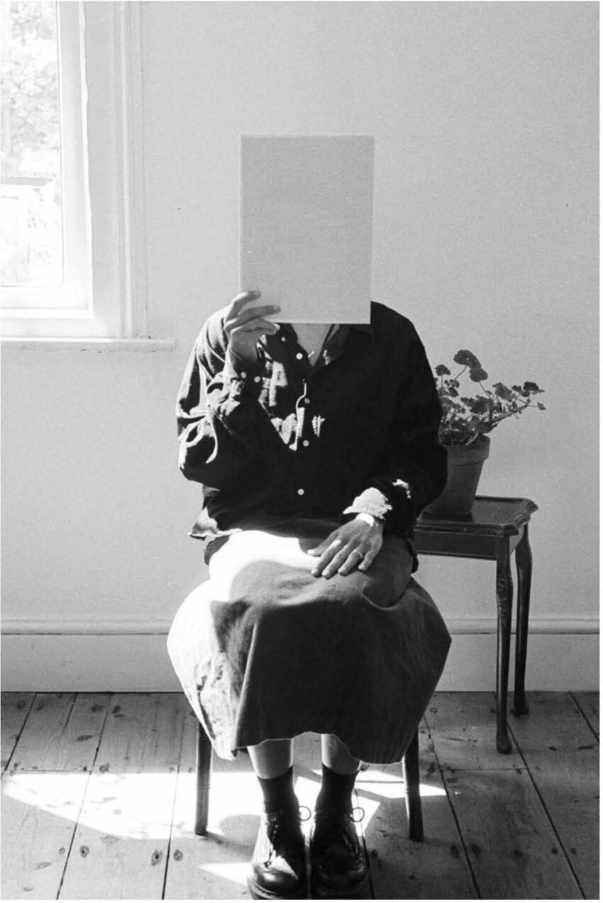 A woman sits before a potted plant, holding up a blank piece of paper which obscures her face